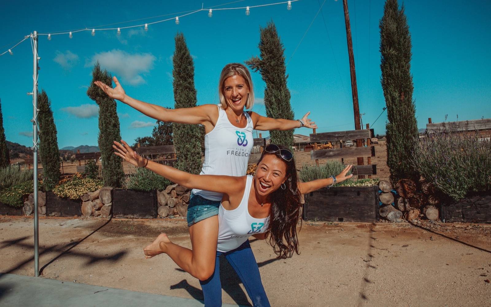 Two women piggy backing each other wearing Flow 2 Freedom Apparel White Aspire bamboo logo tank top